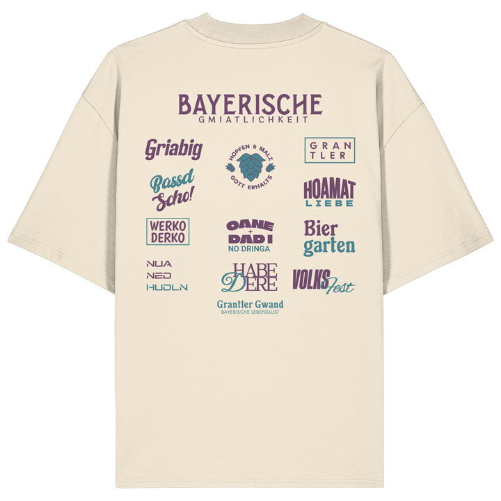 Beiges Oversize-Shirt mit großem lila Backprint „Bayerische Gmiatlichkeit“ und typischen bayerischen Begriffen wie Hoamatliebe, Biergarten, Volksfest, Grantler, Hopfen &amp; Malz und Griabig. Unisex Premium-Shirt aus Bio-Baumwolle im modernen Streetwear-Style. Designed und bedruckt in Bayern von Grantler Gwand.